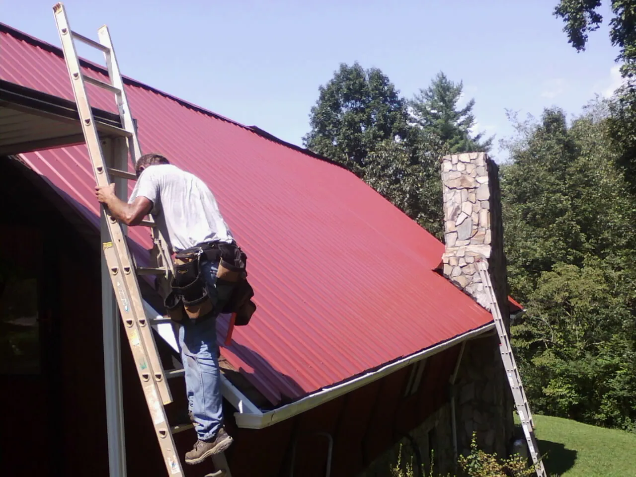 Roofer on a ladder beside a red metal roof and stone chimney — Soffit & Fascia Repair by Roofing Brothers in Benzonia